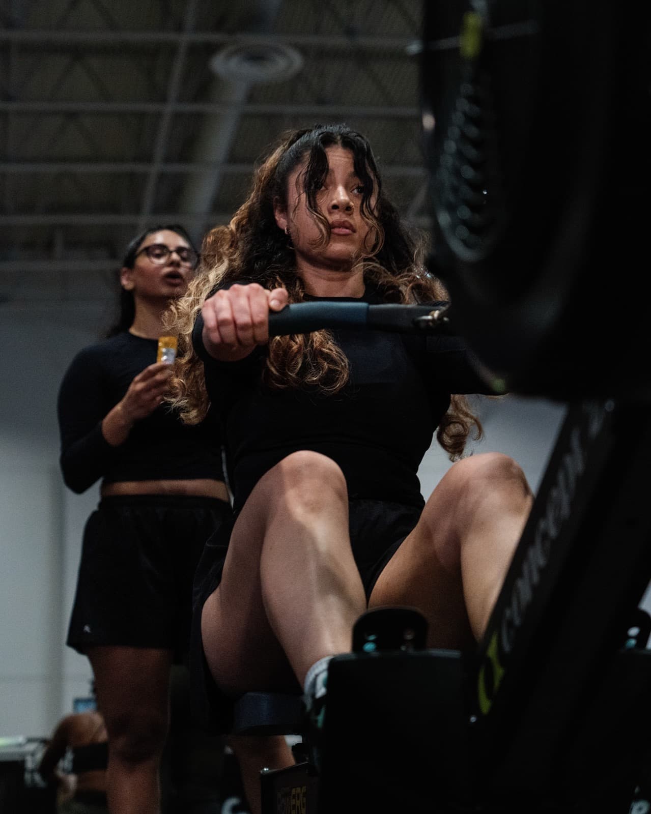An athlete driving through the rowing stroke, a teammate holding a gel behind her