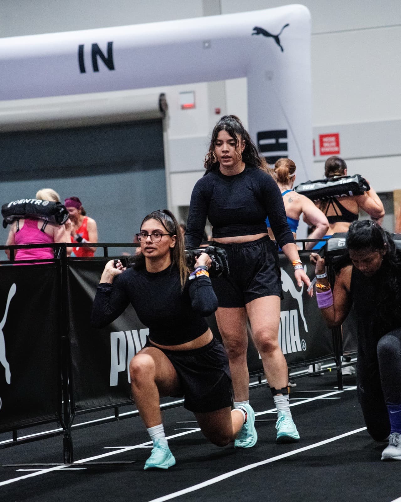 Two HYROX athletes in black carrying sandbags through lunges on the race track