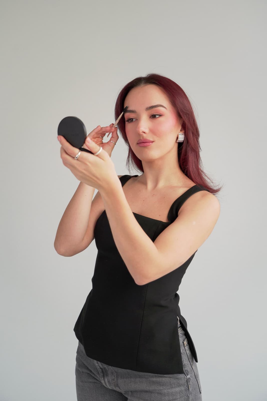 Brushing her brows in a compact mirror against a grey backdrop